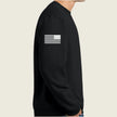 A person wearing a black crewneck sweatshirt, shown from the side. The sweatshirt features a white distressed American flag graphic printed on the upper left sleeve.
