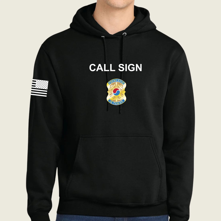 A black pullover hoodie worn by a model, featuring the words 'CALL SIGN' in white across the chest, a gold and blue army badge graphic below it, and a black-and-white American flag on the right sleeve.
