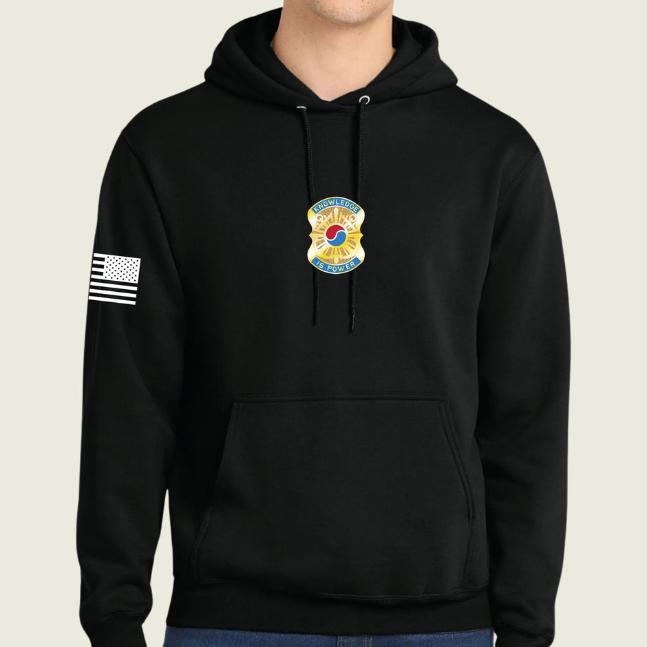 A black pullover hoodie worn by a model, featuring a gold and blue army badge graphic on the center chest and a black-and-white American flag graphic on the right sleeve, set against a solid light beige background.