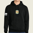 A black pullover hoodie worn by a model, featuring a gold and blue army badge graphic on the center chest and a black-and-white American flag graphic on the right sleeve, set against a solid light beige background.