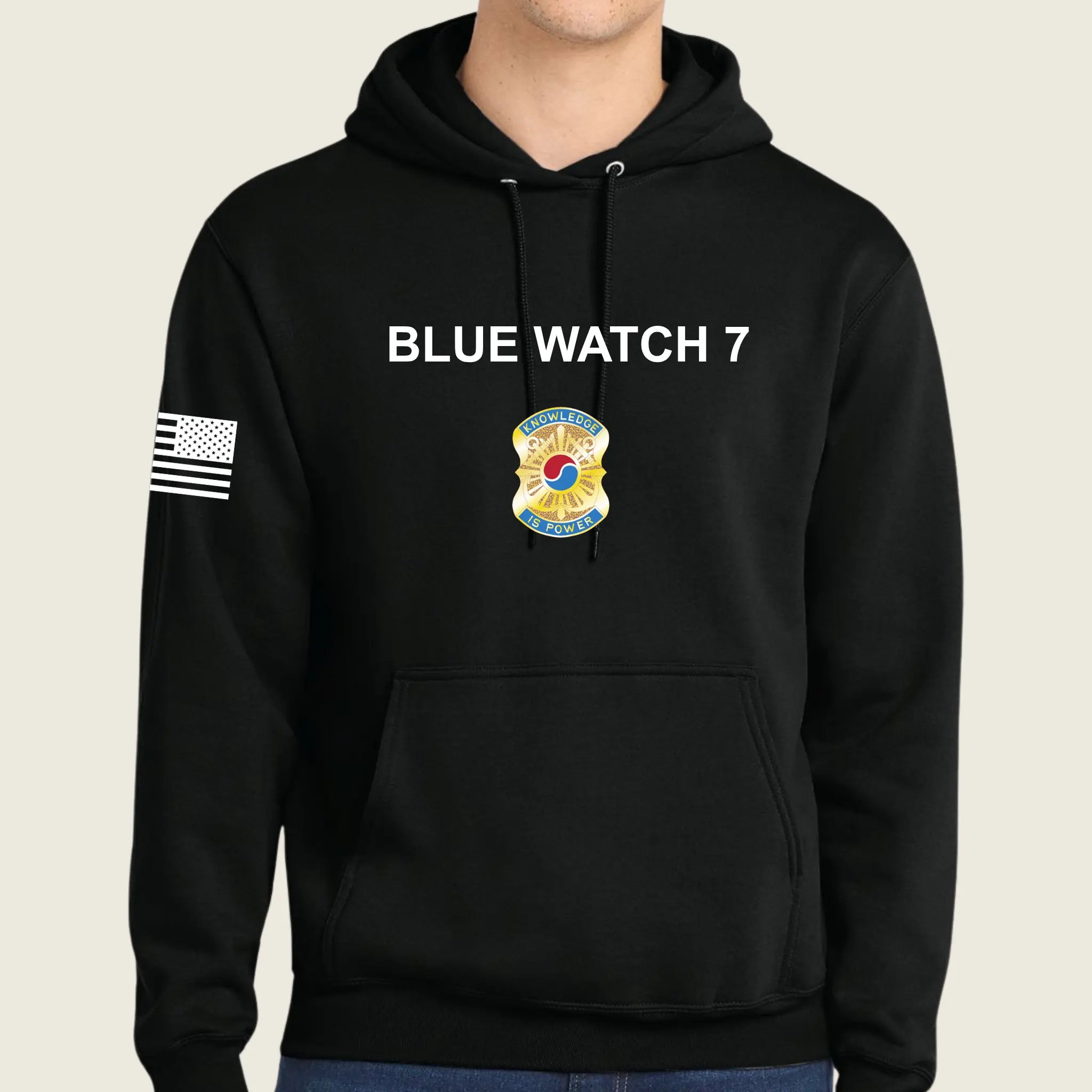 A black pullover hoodie worn by a model, featuring 'BLUE WATCH 7' in white text on the chest, a army graphic below it, and a black-and-white American flag patch on the right sleeve.