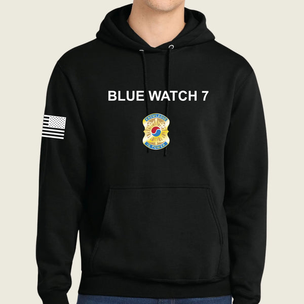 A black pullover hoodie worn by a model, featuring 'BLUE WATCH 7' in white text on the chest, a army graphic below it, and a black-and-white American flag patch on the right sleeve.