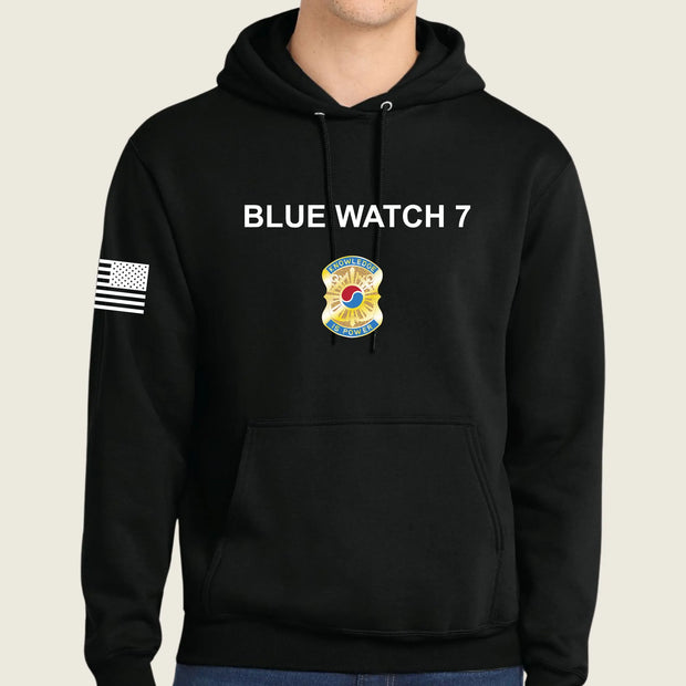 A black pullover hoodie worn by a model, featuring 'BLUE WATCH 7' in white text on the chest, a army graphic below it, and a black-and-white American flag patch on the right sleeve.