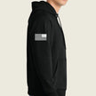 A man wearing a black pullover hoodie with a white distressed American flag graphic printed on the left sleeve, shown in a side profile view against a light beige background.