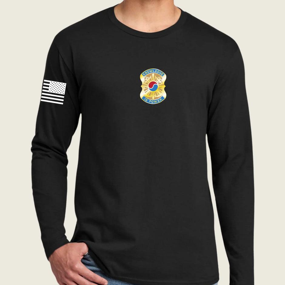 A man wearing a black long-sleeve t-shirt with a black-and-white American flag patch on the right sleeve and a yellow, blue, and army emblem on the chest, set against a light beige background.