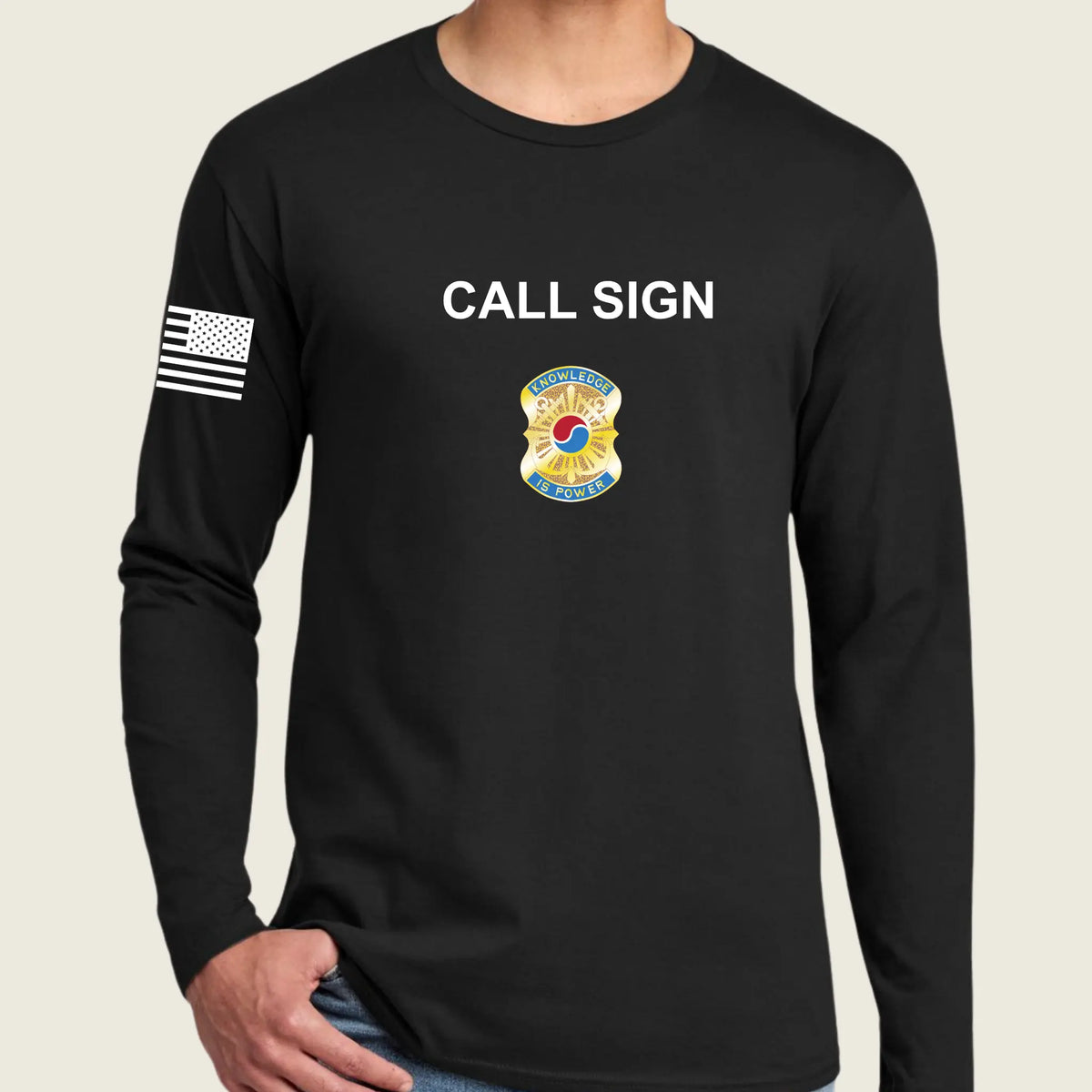 A man wearing a black long-sleeve t-shirt with 'CALL SIGN' printed in white on the chest, a army badge graphic below it, and a black-and-white American flag on the right sleeve, against a light beige background.