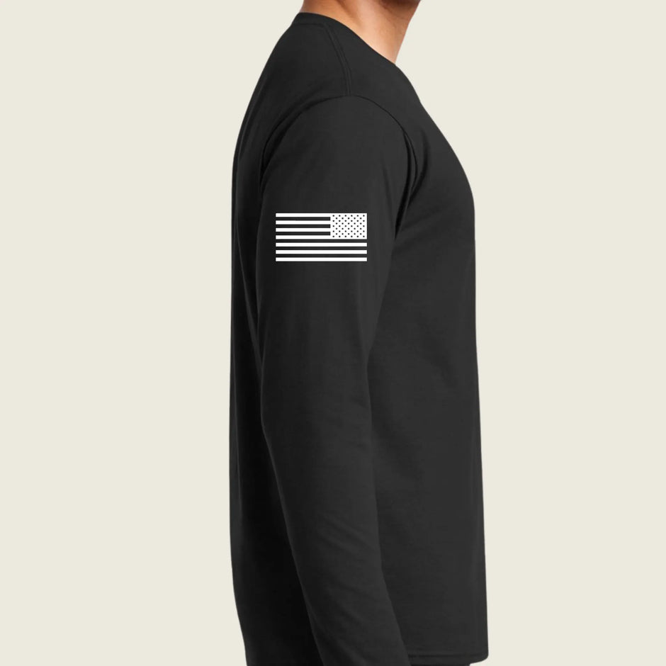A man wearing a black long-sleeve shirt, shown from the side against a light beige background. The shirt features a white distressed American flag graphic printed on the upper left sleeve.