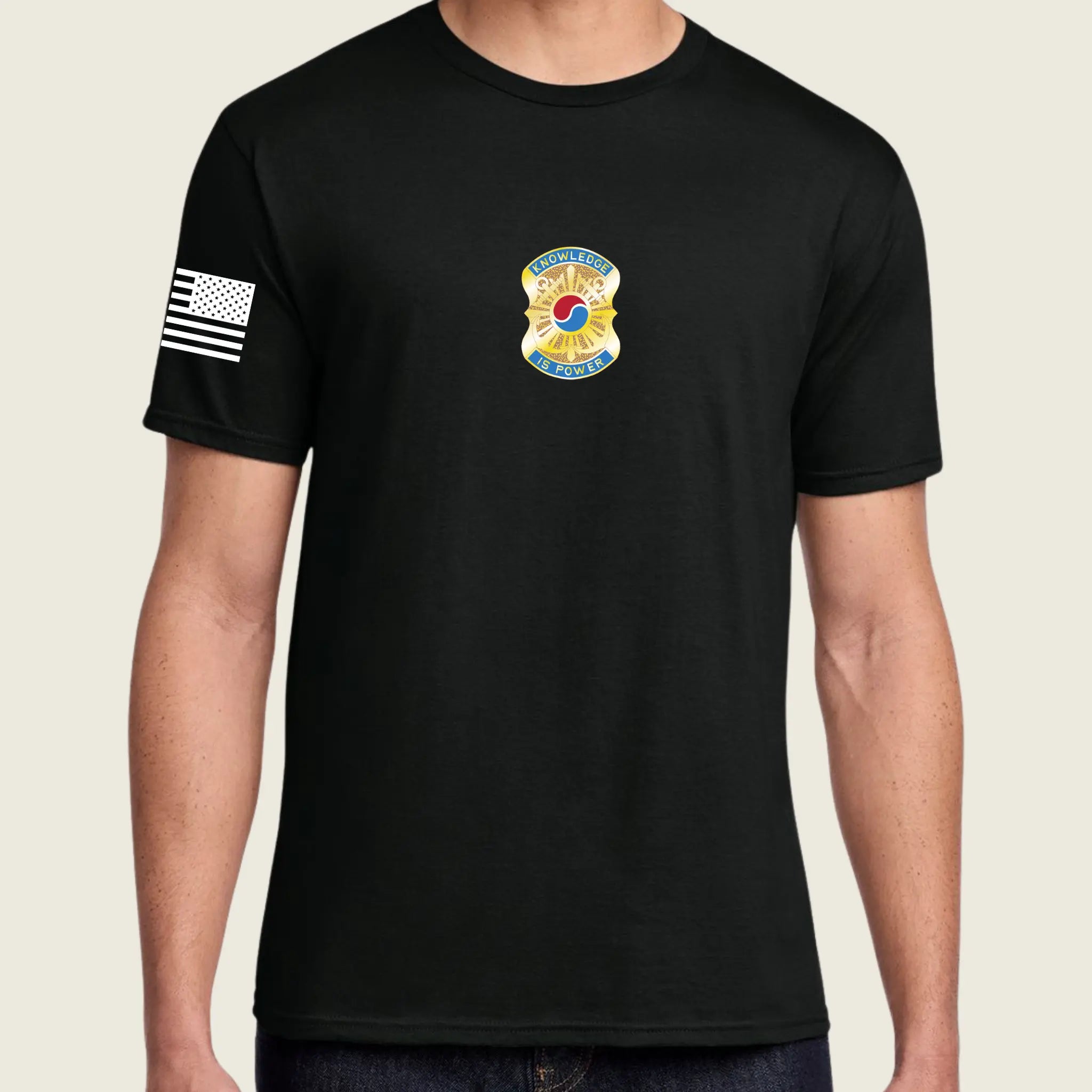 A man wearing a black short-sleeve t-shirt with a yellow and blue badge on the left chest and a black and white American flag graphic on the right sleeve, set against a solid light beige background.