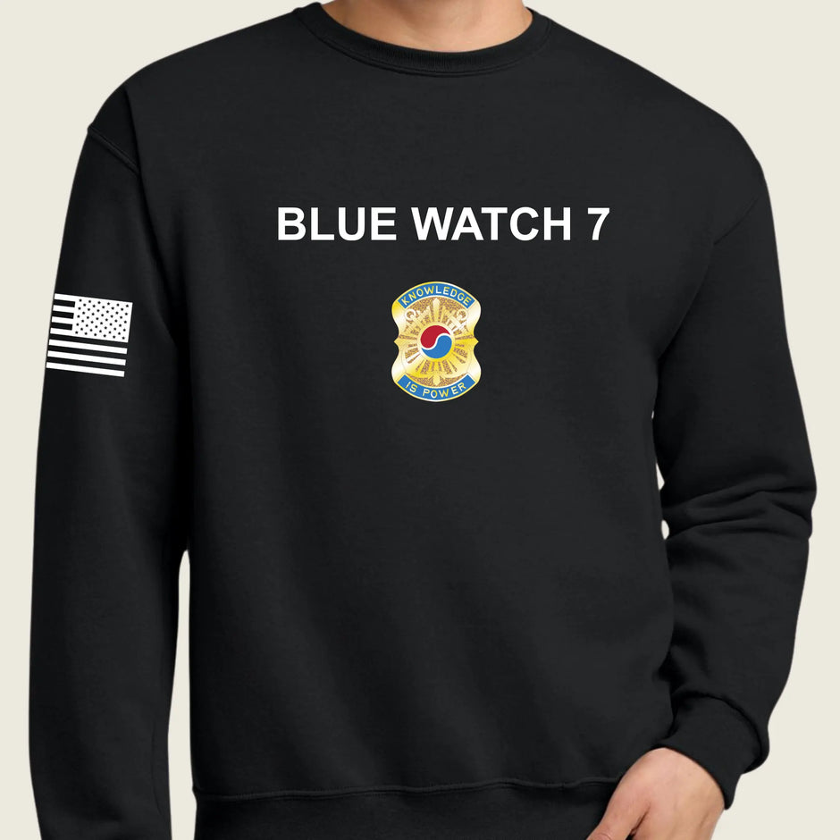 A black crewneck sweatshirt featuring 'BLUE WATCH 7' in white text on the chest, a gold and blue badge emblem below it, and a black-and-white American flag on the right sleeve, shown on a model.
