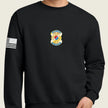 A black crewneck sweatshirt featuring a gold and blue US Army insignia on the chest and a black-and-white American flag patch on the right sleeve, shown on a model.