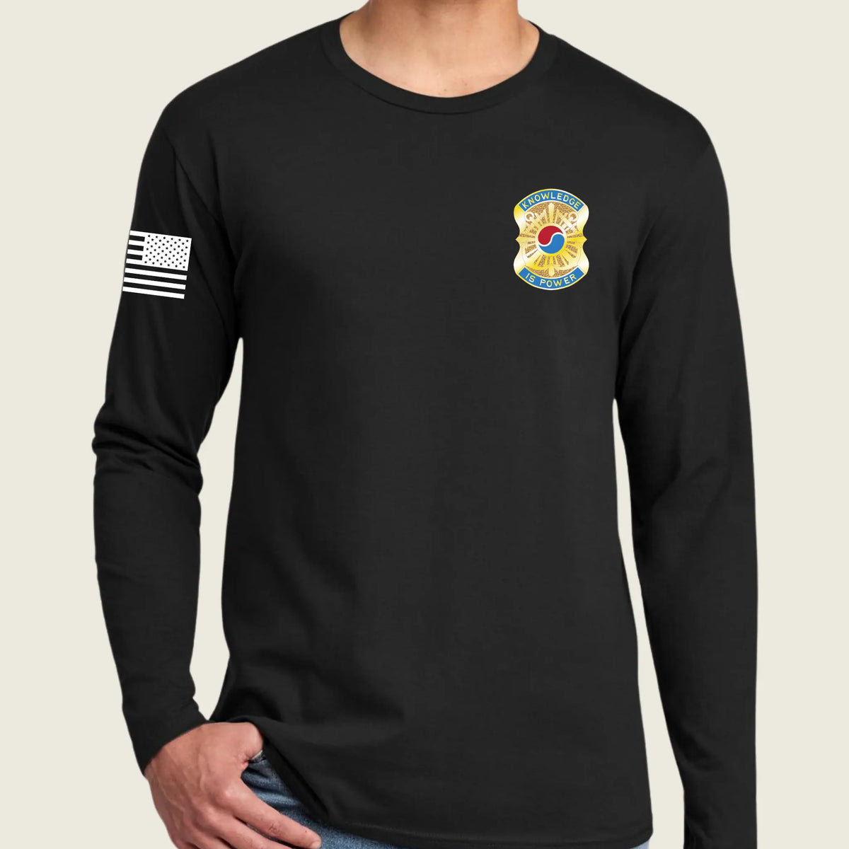 A man wearing a black long-sleeve t-shirt with a US Army insignia on the left chest and a black-and-white American flag on the right sleeve, shown against a light beige background.