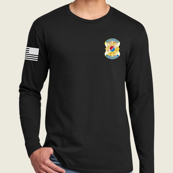 A man wearing a black long-sleeve t-shirt with a US Army insignia on the left chest and a black-and-white American flag on the right sleeve, shown against a light beige background.