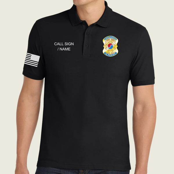 A man wearing a black short-sleeve polo shirt with customizable white text on the left chest reading 'CALL SIGN / NAME' and a white American flag patch on the right sleeve. The left chest features a gold and blue US Army emblem.