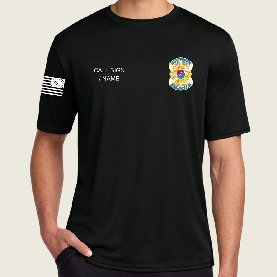 A man wearing a black short-sleeve t-shirt with customizable white text on the left chest, a black and white American flag graphic on the right sleeve, and a yellow and blue badge on the left chest.