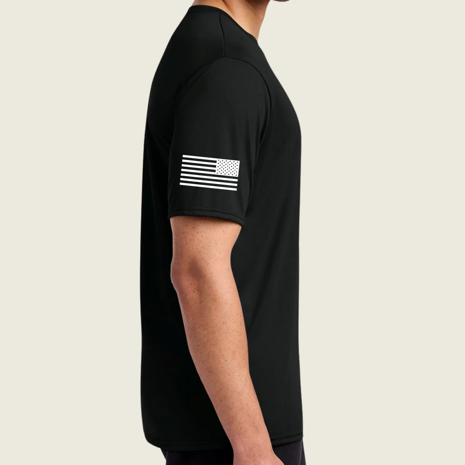 A man wearing a black short-sleeve athletic t-shirt shown from the side profile. The shirt features a distressed white American flag graphic printed on the upper left sleeve, set against a solid light beige background.