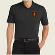 A man wearing a black short-sleeve polo shirt with a gold, red, and black military-style crest patch on the left chest, paired with black trousers and a black belt, against a light beige background.