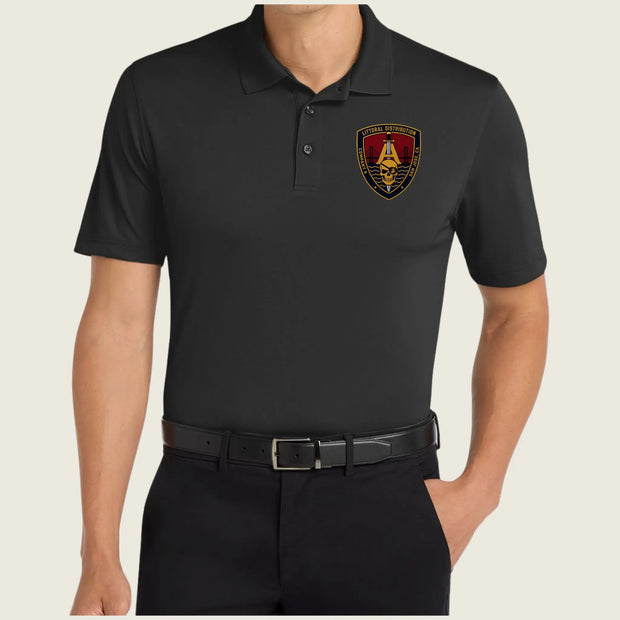 A man wearing a black short-sleeve polo shirt with a gold, red, and black military-style crest patch on the left chest, paired with black trousers and a black belt, against a light beige background.