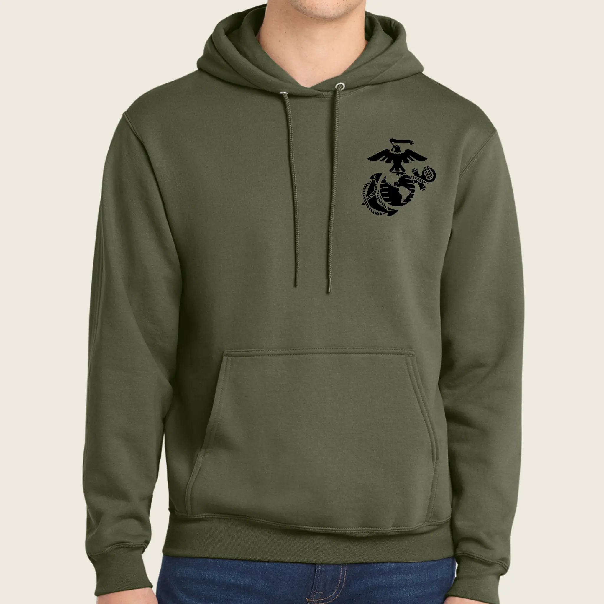 An olive green pullover hoodie with a black US Marine Corps Eagle, Globe, and Anchor emblem printed on the left chest, shown on a model wearing blue jeans against a light beige background.