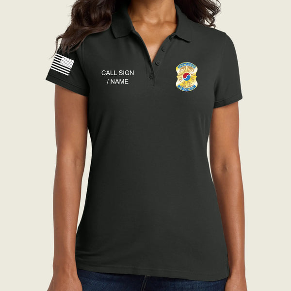 A woman wearing a black polo shirt with a custom printed badge on the left chest, a white text placeholder on the right chest, and a black-and-white American flag patch on the sleeve, shown against a light beige background.