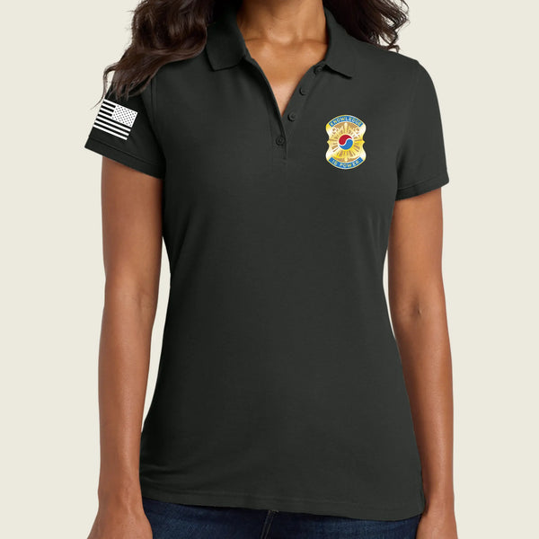 A woman wearing a black short-sleeve polo shirt with a yellow and blue badge on the left chest and a black-and-white American flag patch on the right sleeve, shown against a light beige background.
