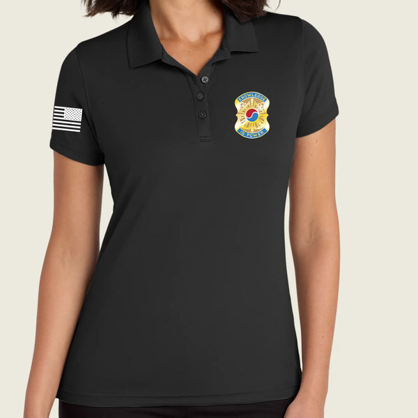 A woman wearing a black short-sleeve polo shirt with a yellow, blue, and red  badge graphic on the right chest, plus a white American flag print on the sleeve.